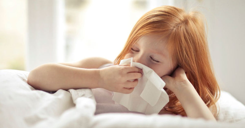 A young girl blows her nose while laying in bed.