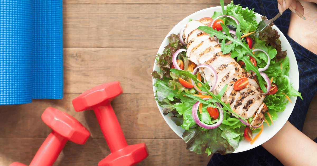 A bowl of salad with grilled chicken strips next to a blue yoga mat and red hand weights.