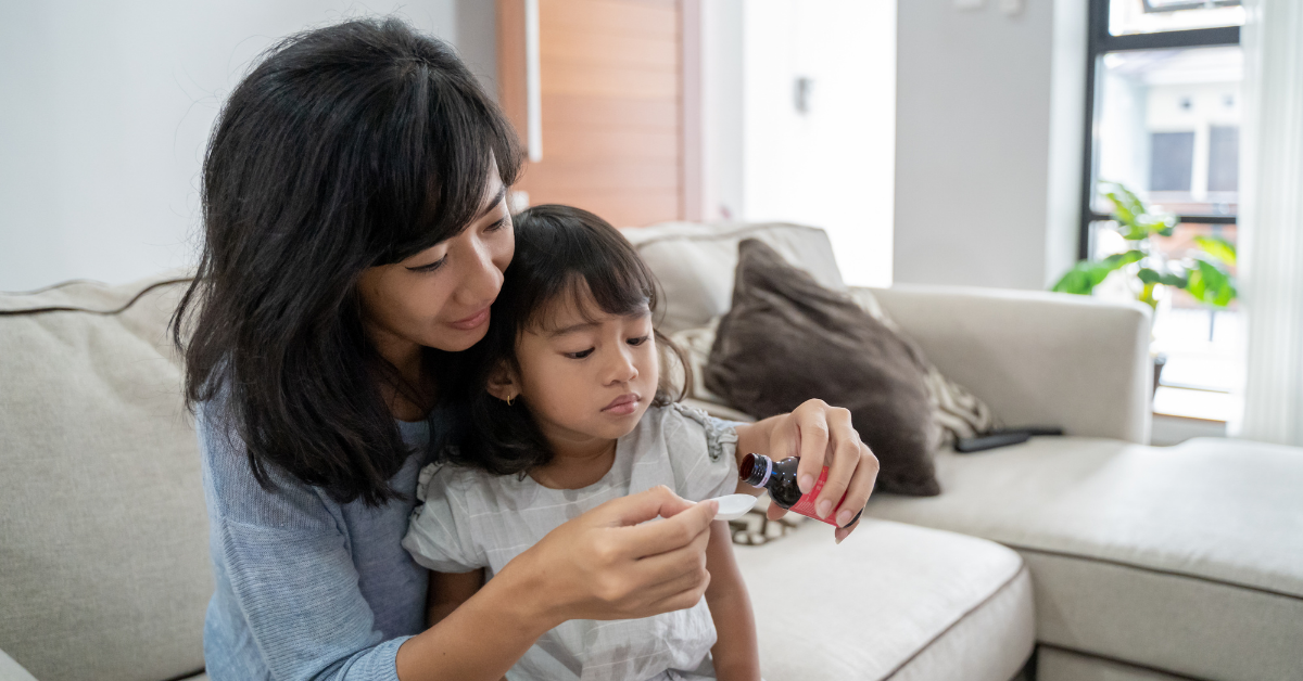 A mother pours a supplement into a small cup for the child sitting on her lap. 