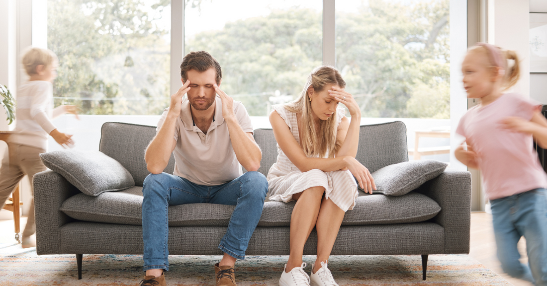 A worn-out mother and father sit on a couch with their hands on their heads while two children run around the couch.