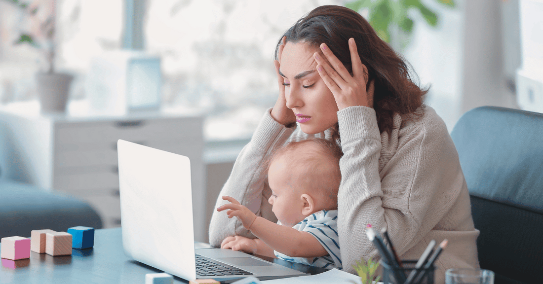 A stressed mom sits at her computer with an baby on her lap.