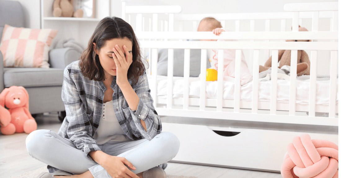 An exhausted mother sits on the floor while her baby plays in a crib.