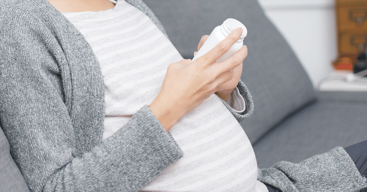 A pregnant woman looks at a supplement bottle while sitting in a gray couch. 