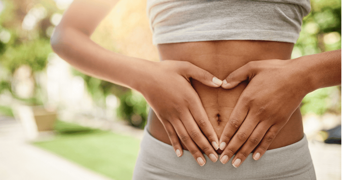 A woman standing outdoors in exercise clothes rests her hands rest on her belly.