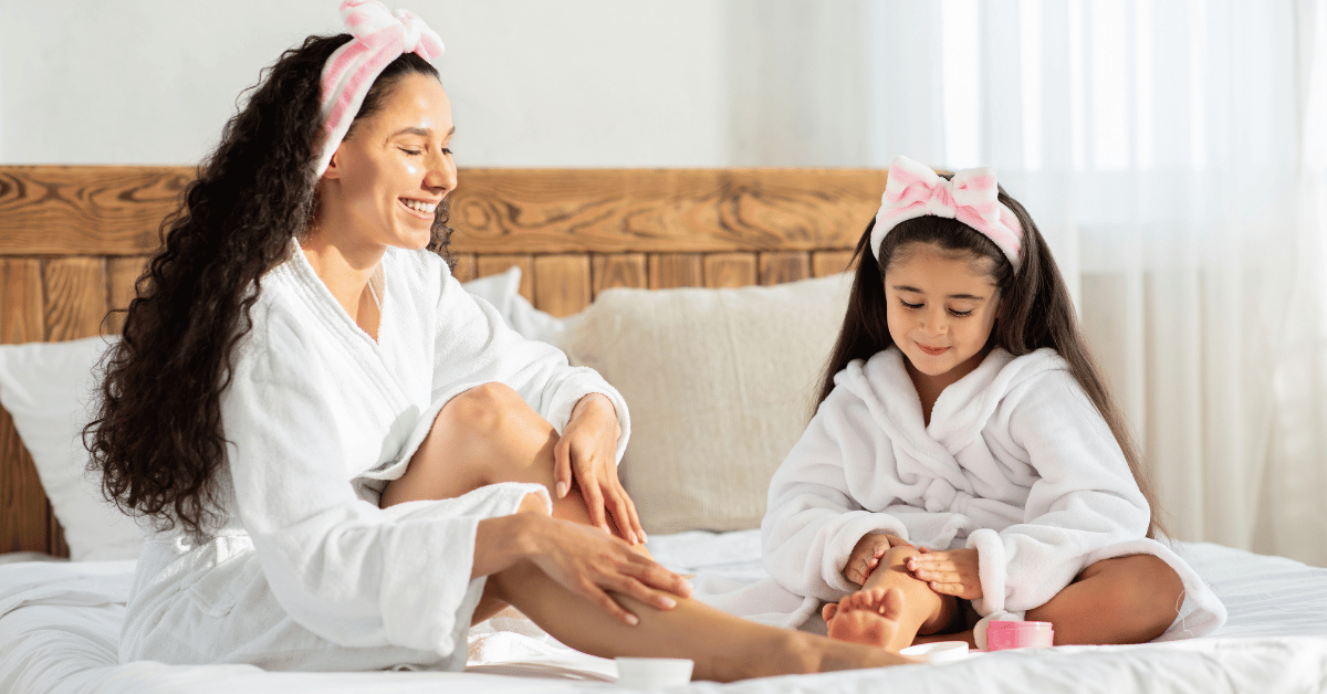A mother and daughter wearing white robes apply lotion to their legs.