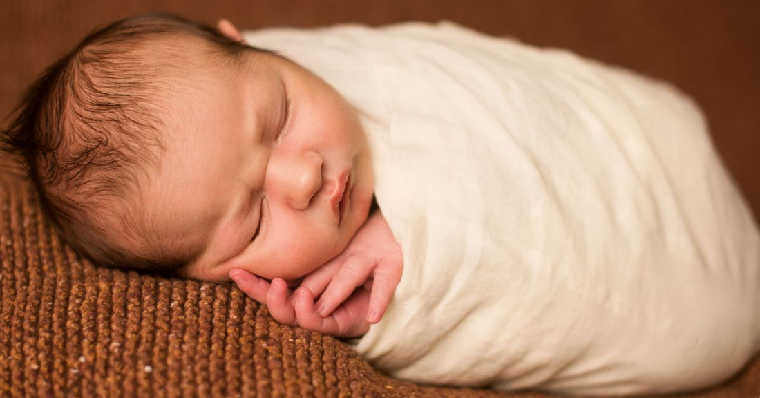 A newborn is swaddled in a white cloth laying on a brown blanket.
