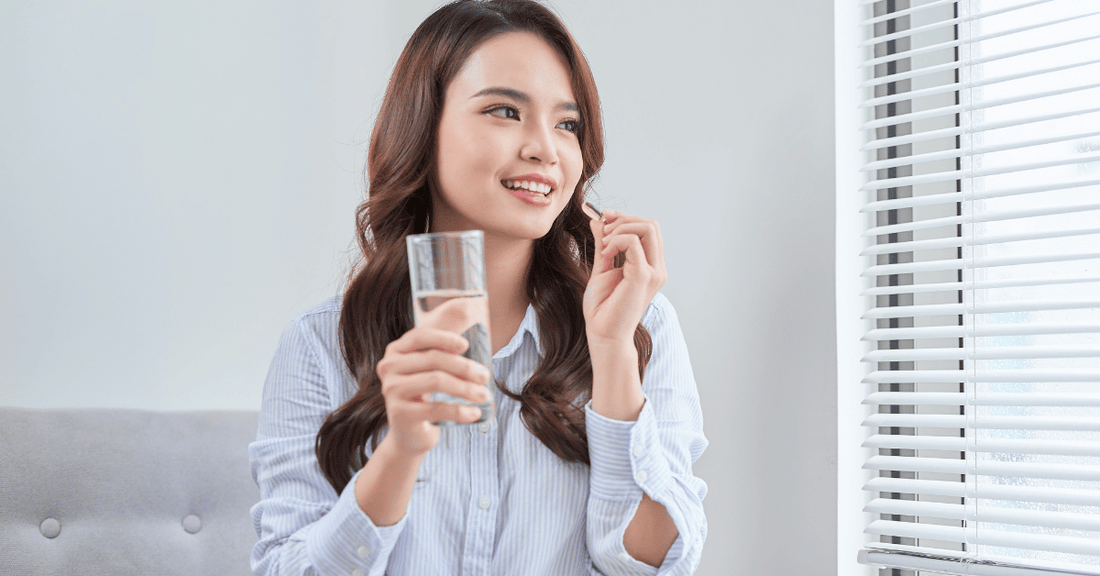 A woman holds a glass of water and a vitamin while looking out the window.